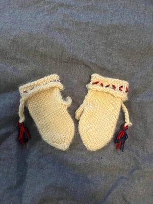 Handstickade vita ullvantar med tofsar (barn) - Säljer ett par handstickade vita vantar i ull smyckade med flätad snodd och tofsar i voice navy och rött. Vantarna har ett dekorativt broderi i rött och blått runt mudden och känns riktigt mysiga för kalla dagar.