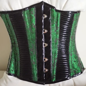 Underbystkorsett 22 med lack detaljer och grön brokad - Säljer en unik underbystkorsett i grön brokad med glansiga svarta lack paneler. Framsidan har metallknäppning och baksidan har justerbar snörning för perfekt passform. Korsett storleken är 22 vilket betyder att du kan snöra ned din midja till 55cm och få en dramatisk timglasmidja. Passa bra till så väl daglig användning som fest. Köpt hos Corset-Story UK för 900kr. 