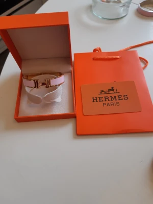 Hermes armband  - Bilderna visar ett armband från Hermès i rosa och guld med tillhörande ask och påse, 