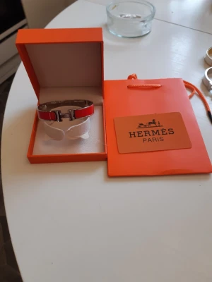 Hermes armband  - Bilderna visar ett Hermès-armband i röd och silverfärg med tillhörande ask och påse,