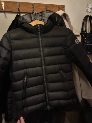 Svart pufferjacka från Colmar - Säljer en svart pufferjacka från Colmar med huva och två dragkedjeförsedda fickor framtill. Jackan har en quiltad design och en snygg logga på ärmen. Perfekt för kalla dagar och riktigt skön att bära. Storlek 40 i italiensk storlek motsvarar XS