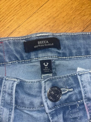 True Religion Becca Bootcut Jeans ljusblå - Ljusblå jeans från True Religion, modell Becca Mid Rise Bootcut. Klassisk femficksdesign med snygga detaljer och logoknappar, samt ficklock bak. Jeansen har bootcut-ben och är tillverkade i jeansmaterial.