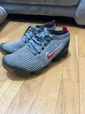 Nike Vapormax skor  - Säljer ett par grå Nike Vapormax skor med stickad ovandel och röda detaljer. Skorna har en tydlig röd Nike logo på sidan, svart sula med synliga luftbubblor och snörning. Perfekta för dig som vill ha något bekvämt och snyggt ingen av sulorna är sönder helt ny annars så kan du returnera skorna direkt. Skorna är äkta 100%. 