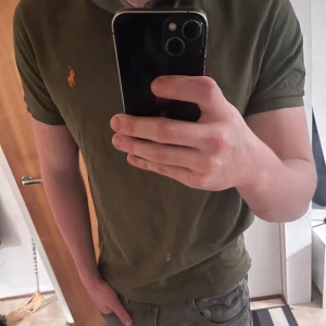 Olivgrön piké från Polo Ralph Lauren - Säljer en olivgrön pikétröja från Polo Ralph Lauren i custom slim fit. Klassisk orange logga på bröstet och mjukt bomullsmaterial. Perfekt för dig som gillar stilrena och tidlösa plagg med snygg passform.