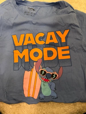 Blå Disney t-shirt med Stitch-print - Säljer en blå t-shirt från Disney med stort tryck av Stitch i solglasögon och texten 'VACAY MODE' i orange. T-shirten har rund hals och korta ärmar, perfekt för dig som gillar coola prints och tecknade karaktärer.