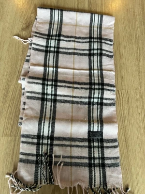 Rutig halsduk från Burberrys - Säljer en klassisk rutig halsduk från Burberrys i ljusrosa, svart och beige. Halsduken är tillverkad i 100% lammull och har fransar i ändarna. Perfekt accessoar för att lyfta din outfit med en tidlös brittisk vibe.