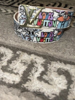 Färgglad läder  bälte - Säljer ett unikt  bälte med färgglatt tryck och tecknade figurer. Bältet har en stor guldfärgad -logga som spänne och bakgrunden är vit med inslag av blått, orange och svart. Perfekt för dig som vill sticka ut med accessoarer.