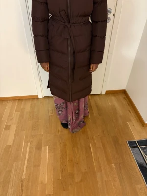 Brun lång pufferjacka med bälte - Säljer en lång, mörkbrun pufferjacka med bälte i midjan och dragkedja framtill. Jackan har långa ärmar och quiltad design som ger en riktigt mysig känsla. Perfekt för kalla dagar när du vill hålla dig varm och ändå se snygg ut. Helt ny knappt använd 