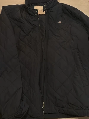 Svart quiltad jacka från Gant - Säljer en svart quiltad jacka från Gant i storlek 2XL. Jackan har hög krage med tryckknappar, dragkedja framtill och två sidofickor. Klassisk Gant-logga broderad på bröstet. Perfekt för kyliga dagar och enkel att matcha med det mesta.
