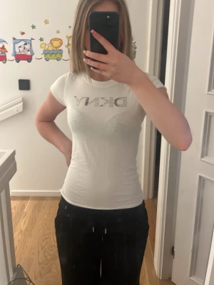 Vit DKNY t-shirt - Säljer en vit t-shirt från DKNY, framsidan har ett DKNY-tryck i silverfärgade nitar som ger en snygg touch. Den är väldigt mjuk att ha på sig och passar perfekt till jeans eller shorts för en clean look! Den är storlek L men som man ser sitter den rätt så tight (jag brukar ha XS). Jag älskar tröjan men behöver lite extra pengar just nu! Skriv för mer bilder eller andra frågor! 💗