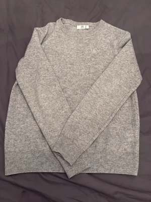 Grå stickad tröja från Bivaldi - Mysig grå stickad tröja från Bivaldi med diskret logga broderad på bröstet. Klassisk rund halsringning och ribbade muddar vid ärmslut och nederkant. Perfekt för lager på lager under kyliga dagar.