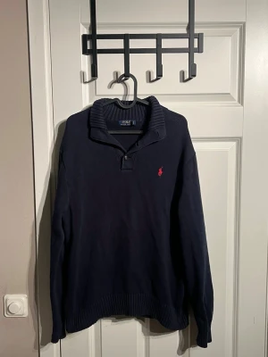 Mörkblå stickad tröja Polo Ralph Lauren - Mörkblå stickad tröja från Polo Ralph Lauren med ribbad krage och knappdetalj vid halsen. Klassisk röd logga broderad på bröstet. Mer frågor eller bilder är det bara att fråga!