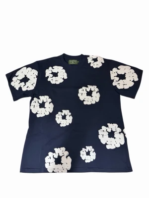 Blå blommig t-shirt från Denim Tears - Mörkblå t-shirt från Denim Tears med stora vita blommiga tryck över hela plagget. Klassisk rund hals och korta ärmar. Tillverkad i mjuk bomull som känns skön mot huden. Perfekt för dig som vill sticka ut med en unik och trendig look.