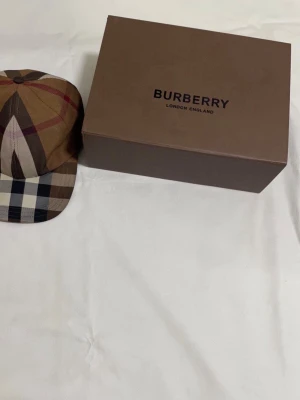  Burberry keps i bomull - Säljer en klassisk Burberry keps i storlek M med ikoniskt rutigt mönster i beige, brunt, svart och vitt. Kepsen är tillverkad i 100% bomull och har justerbart spänne baktill med Burberry-logga. Perfekt accessoar för att lyfta din outfit.