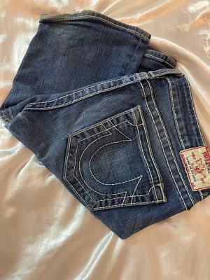 True Religion blå bootcut jeans - Säljer ett par klassiska blå jeans från True Religion med tydliga kontrastsömmar och ikoniska bakfickor med vit brodyr. Jeansen har bootcut-modell och är tillverkade i mjukt jeansmaterial. Märkespatch i rött och vitt på linningen bak. Säljer för att de är för långa för mig i benen, de finns tydliga tecken på att de tidigare har sytts igen ( inte av mig) , där av de lägre priset men inget som jag uppfattar som tydligen när man har på sig dom, finns tydligare bilder på de om man vill se hör av er!