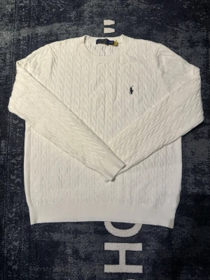 Vit kabelstickad tröja Polo Ralph Lauren - Säljer en klassisk vit kabelstickad tröja från Polo Ralph Lauren. Tröjan har rund hals, lång ärm och den ikoniska broderade loggan på bröstet. Perfekt för dig som gillar stilrena och tidlösa plagg.