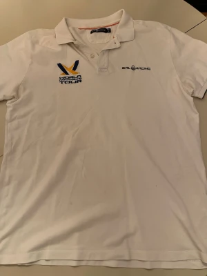 Vit piké från Sail Racing XL - Klassisk vit pikétröja från Sail Racing i storlek XL. Tröjan har broderade loggor på bröstet, inklusive World Match Racing Tour och Sail Racing. Tillverkad i bomull med krage och knappar framtill. Perfekt för en sportig och avslappnad stil.