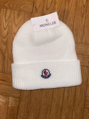 Vit ribbstickad mössa från Moncler - Säljer en stilren vit ribbstickad mössa från Moncler med uppvikt kant och klassisk Moncler-logga framtill. Perfekt accessoar för kyliga dagar och enkel att matcha med olika outfits.