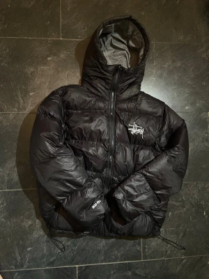 Svart pufferjacka från Stüssy - Säljer en svart pufferjacka från Stüssy med huva och dragkedja framtill. Jackan har Stüssy-logga broderad på bröstet och justerbara snören nedtill. Perfekt för kalla dagar och riktigt snygg streetwear-vibe.