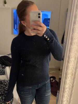 Mörkblå tröja Holly & Whyte  - Mörkblå långärmad tröja från Holly & Whyte med snygga guldfärgade knappar vid ärmsluten. Tröjan har rund halsringning och är i mjukt material som känns skönt mot huden. Perfekt till jeans eller kjol för en clean look.