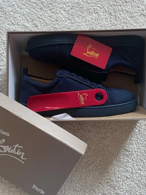 Mörkblå mocka sneakers från Louboutin - Snygga mörkblå sneakers från Christian Louboutin i mjuk mocka med klassisk röd sula. Skorna har rund tå, snörning och diskreta detaljer. Perfekta för dig som vill ha en clean och exklusiv look med en touch av lyx.