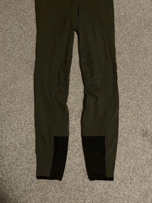 Mörkgröna ridbyxor CRW strl 34 - Säljer ett par mörkgröna ridbyxor från CRW med silikon-grepp på insidan av benen och svarta detaljer vid bensluten. Byxorna har hög midja, dragkedja och dubbla knappar framtill. Snygga och funktionella för ridning. har aldrig använt dom🥰