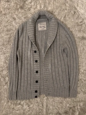 Grå stickad kofta  - Grå ribbstickad kofta med bred krage och svarta knappar framtill. Koftan har lång ärm och är gjord i mjukt stickat material, perfekt för kyliga dagar. Klassisk och enkel design som är lätt att matcha. Kontakta gärna om ni har några frågor eller funderingar gällande pris eller plagg