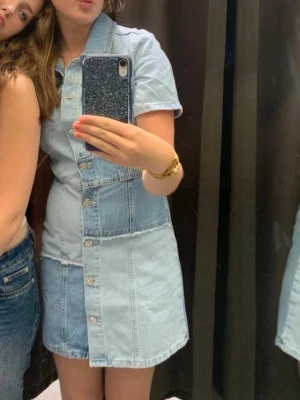 Patchwork jeansklänning med knappar - Säljer en kort jeansklänning i patchwork-stil med ljusblå och mellanblå denim. Klänningen har korta ärmar, krage och knappar hela vägen framtill. Snygga råa kanter mellan denimlapparna ger en cool vibe. Perfekt för dig som gillar unik stil!