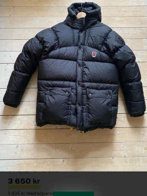 Svart dunjacka från Fjällräven st SMALL - Säljer en svart, riktigt fet dunjacka från Fjällräven med hög krage och två stora fickor framtill. Jackan har gula dragskor och klassisk Fjällräven-logga på bröstet. Perfekt för kalla dagar när du vill hålla värmen och ändå se clean ut.
