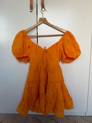 Orange kortklänning från Zara - Superfin orange klänning från Zara med puffärmar och broderade blommor. Klänningen har omlottkänsla upptill, smock i ryggen och volangdetaljer i kjolen. Perfekt för varma dagar och ger en färgklick till garderoben.
