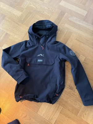 Svart vindjacka från Dope - Säljer en svart vindjacka från Dope med huva och stor magficka med DOPE-logga. Jackan är nästan helt oanvänd och i storlek s säljer även ett par byxor som hör ihop
