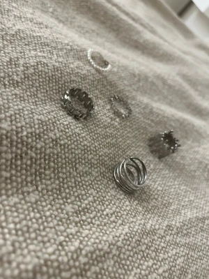 Snygga silvriga ringar i olika modeller - Säljer ett set med fem silvriga ringar i olika coola designer. En ring har ett organiskt, droppformat mönster, en annan består av flera tunna ringar i lager, och en tredje har ett kedjeinspirerat utseende. Perfekta för att mixa och matcha till din stil.