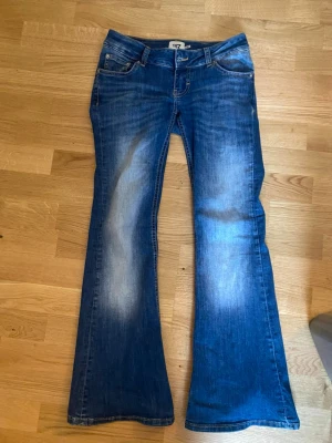 Blå bootcut jeans med slitningar - Säljer ett par blå jeans med bootcut-modell och snygga slitningar på låren. Jeansen har klassisk femficksdesign, låg midja och är tillverkade i ett mjukt denimtyg. Perfekta för dig som gillar retrovibbar och vill ha ett par jeans som sticker ut.