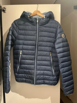 Blå dunjacka från JOTT med huva - Säljer en blå dunjacka från JOTT med huva och dragkedja framtill. Jackan har två sidofickor med dragkedja och ett broderat JOTT-märke på ärmen. Lättviktigt material och quiltad design gör den perfekt för kyliga dagar.