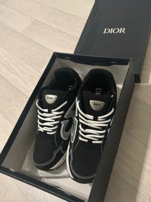 Dior b30 svarta. - Säljer ett par svarta Dior sneakers med vita och silverfärgade detaljer. Skorna har tydliga CD-loggor på sidan och hälen, samt vita snören och en chunky vit sula. Materialmix av mesh och syntet ger en sportig vibe. Perfekta för dig som vill ha en lyxig touch på din streetwear.