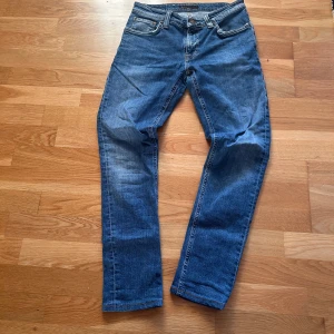 Blå jeans från Nudie Jeans - Säljer ett par klassiska blå jeans från Nudie Jeans med snygga slitningar och kontrastsömmar. Modellen har raka ben, fem fickor och läderpatch med logga bak. Jeansen är tillverkade i slitstarkt bomullsmaterial och har en tidlös look. Dem är rätt så tajta