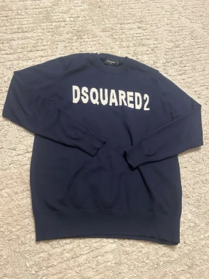 Marinblå tröja från Dsquared2 - Säljer en marinblå tröja från Dsquared2 med vit logga framtill. Tröjan har rund halsringning, ribbade muddar och är långärmad. Perfekt för dig som gillar stilrena plagg med tydlig branding.