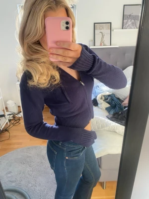 Mörkblå croppad hoodie med dragkedja - Säljer en mörkblå croppad hoodie med dragkedja framtill och huva. Tröjan är stickad med bred ribbad mudd i midjan och vid ärmsluten. Perfekt för dig som gillar en lite mer avslappnad och trendig look.