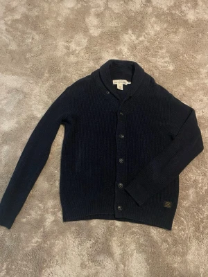 Mörkblå stickad kofta  - Mysig mörkblå stickad kofta från H&M L.O.G.G. med klassisk krage och knappar framtill. Koftan har lång ärm och en liten patch nertill. Perfekt för lager på lager under kyliga dagar. Skickas endast med PostNord. Om annat fraktsätt väljs av Plick, avbryts köpet.