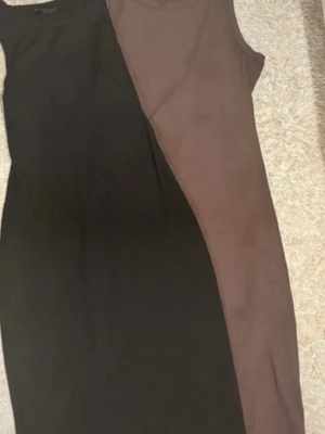 Två klänningar för ett pris - Två snygga långklänningar i svart och brun färg, båda är ribbade och ärmlösa med rund hals. Klänningarna har en slim passform och är gjorda i ett stretchigt material som sitter skönt mot kroppen. Perfekt för dig som gillar enkel och stilren design.