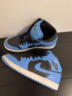 Nike Air Jordan 1 Mid blå/svarta - Nike Air Jordan 1 Mid i blått och svart med klassisk Jordan-logga på sidan. Skorna har snörning, rund tå och platt sula. Tillverkade i skinn och syntetmaterial, med svart innersula och blå detaljer på yttersulan. Perfekt för dig som gillar streetwear och sneakers med ikonisk design.