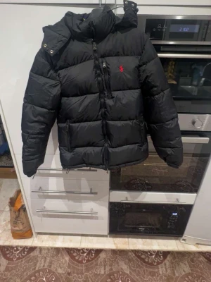 Svart pufferjacka från Polo Ralph Lauren - Säljer en svart pufferjacka från Polo Ralph Lauren med röd broderad logga på bröstet. Jackan har huva, dragkedja framtill och två sidofickor med dragkedja. Insidan är slät och materialet är syntet. Perfekt för kalla dagar. Helt ny passade inte så säljer den istället har aldrig   använt förutom när den  testades hemma har haft den liggandes hemma ett tag men annars helt ny 