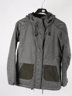 Grå jacka från Fjällräven - Säljer en grå jacka från Fjällräven med huva och svarta snören i midjan och vid halsen. Jackan har stora fickor framtill och knappdetaljer vid ärmsluten. Perfekt för dig som vill ha en stilren och funktionell jacka.