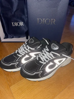 Svarta CD sneakers med vita detaljer - Säljer ett par svarta CD sneakers med vita reflekterande detaljer och vita snören. Skorna har en chunky vit och svart sula, mesh och syntetmaterial på ovansidan samt CD-logga på hälen. Snygg design som sticker ut och levereras med originalkartong och dustbag.