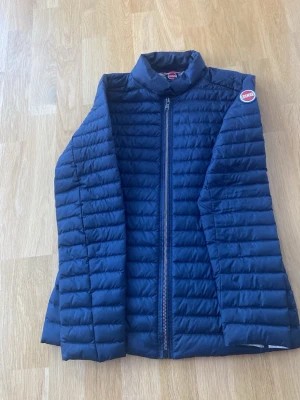 Mörkblå dunväst från Colmar - Säljer en mörkblå dunväst från Colmar med quiltad design och hög krage. Västen har dragkedja framtill och en snygg logga på axeln. Perfekt att slänga över en hoodie eller tröja för extra värme.