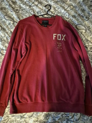Vinröd sweatshirt från FOX, slim fit - Säljer en vinröd sweatshirt från FOX i storlek L. Tröjan har rund hals, tryck med FOX-logga och text på bröstet. Slim fit-modell som sitter snyggt och är tillverkad i mjuk bomull. Perfekt för dig som gillar streetwear och enkel stil.