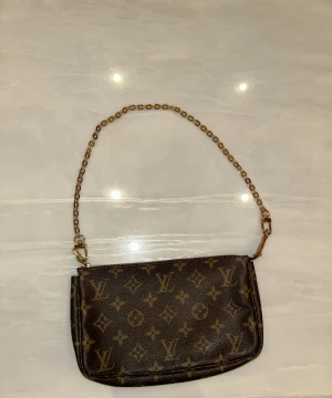 Louis Vuitton monogram axelväska - Säljer en klassisk brun axelväska från Louis Vuitton med ikoniskt monogrammönster och guldfärgade detaljer. Väskan har både en kedjerem och en läderrem som kan bytas ut. Väskan köptes från MerchSweden, läderremet tillhör väskan men kedjeremet köptes separat från de.