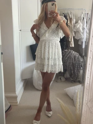 Vit spetsklänning med volanger - Supersöt vit kortklänning i spets med omlott och v-ringning. Klänningen har volangdetaljer, korta ärmar och knyt i ryggen. Perfekt för sommarens alla tillfällen och speciellt student. Materialet är spets med blommigt mönster.🤍