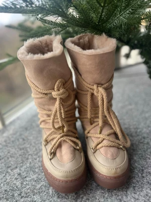 Beige höga Inuikii moon boots med snörning storlek 38 - Säljer ett par beige höga Inuikii boots med grov snörning och fluffigt foder. Skorna har en rund tå, platt sula och är tillverkade i en mix av mocka och skinn. Perfekta för kalla dagar och ger en riktigt mysig vinterlook. Använda 3 gånger och då i nysnö. 