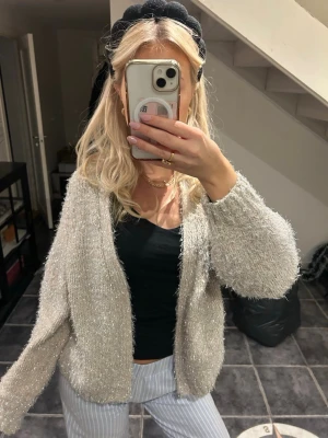 Glittrig beige kofta med lång ärm - Säljer en supermjuk och glittrig beige kofta med lång ärm. Koftan är stickad och har en öppen front utan knappar, vilket ger en chill och avslappnad vibe. Perfekt att slänga över en topp för extra shimmer i vardagen.